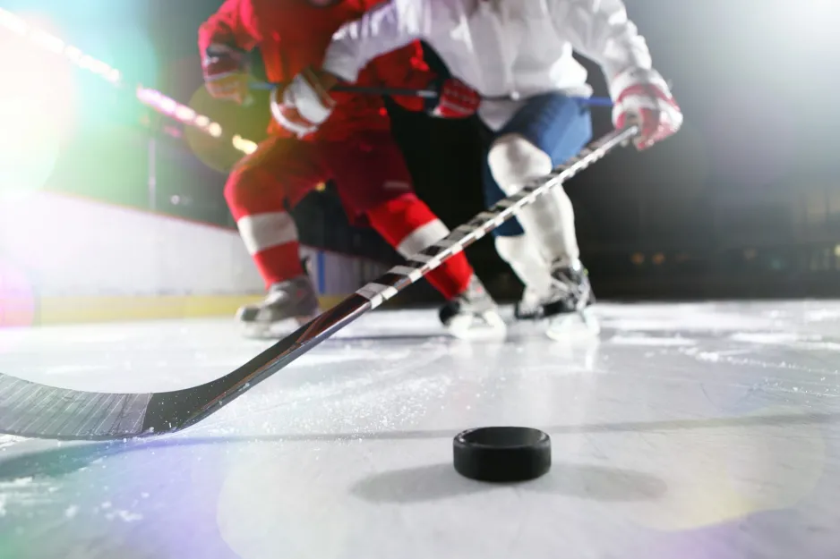 Ice-level image of male ice hockey players in a tackle during a game. The high white socks and dark blue trousers of the hockey player can be seen; there's a hockey stick reaching for the puck. 