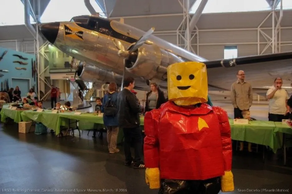A person dressed as a smiling Lego figure at the Museum; we see an aircraft in the background.