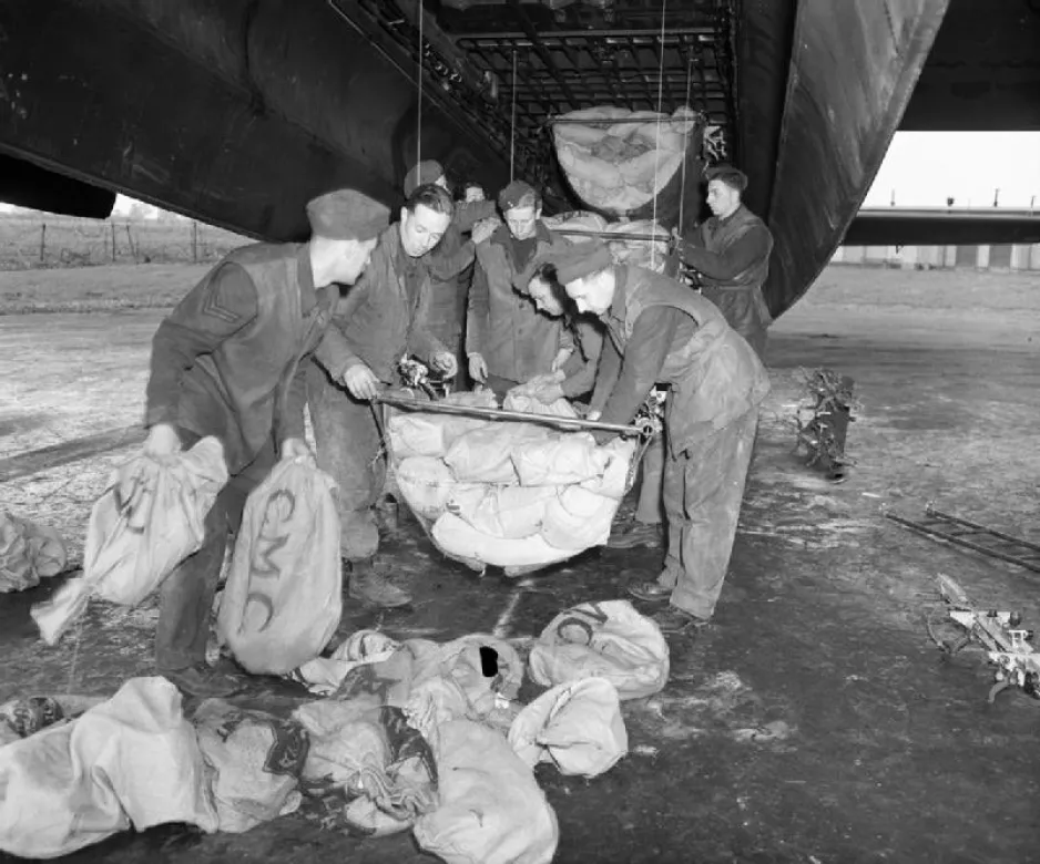 Une image en noir et blanc représente un groupe d'hommes debout sous un Avro Lancaster de la Royal Air Force; ils chargent des sacs d’approvisionnements en tissu dans la soute à bombes. Wikimedia Commons