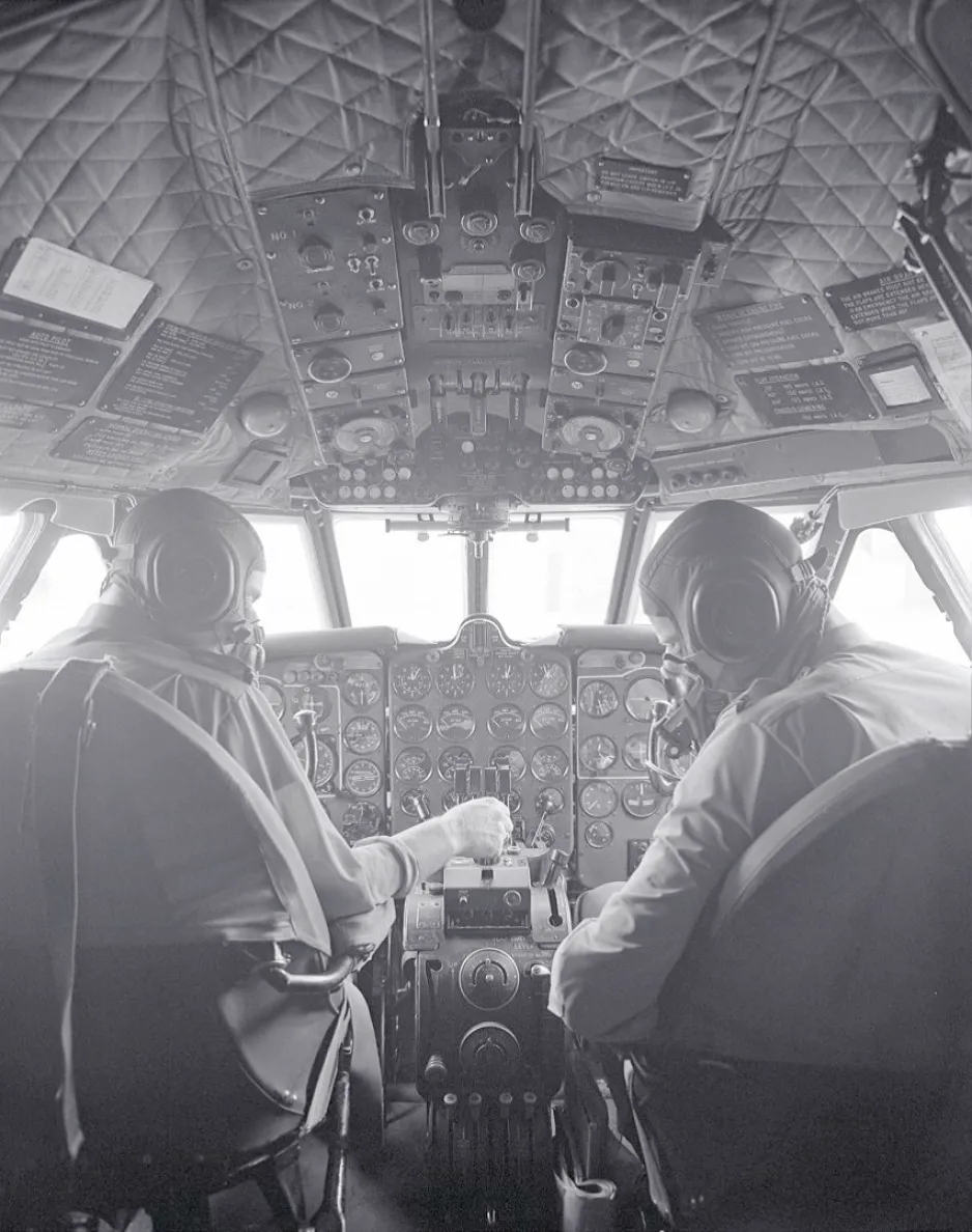 A black-and-white image of the back of two pilots, as they sit at the controls of an aircraft.