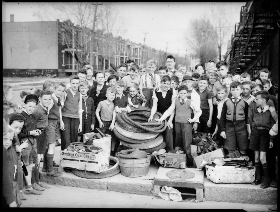 Un grand groupe d’enfants, principalement des jeunes garçons, et un homme, sont rassemblés en demi-cercle sur un trottoir de banlieue, entourant un tas de pneus de voiture et de vélo, et des bottes en caoutchouc qu’ils avaient ramassées.