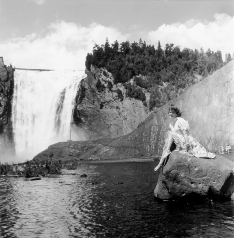 A black-and-white photograph of a scenic view of Montmorency Falls, Quebec. The waterfalls are on the left side of the photo and a woman is posed on a rock on the right side of the image.