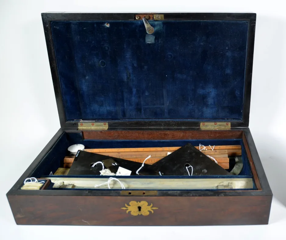 Une jolie mallette en bois, dont l’intérieur est garni d’une doublure bleu royal, est posée sur une table; elle contient d’anciens instruments de dessin. La photo montre l’artefact avant son traitement de conservation.