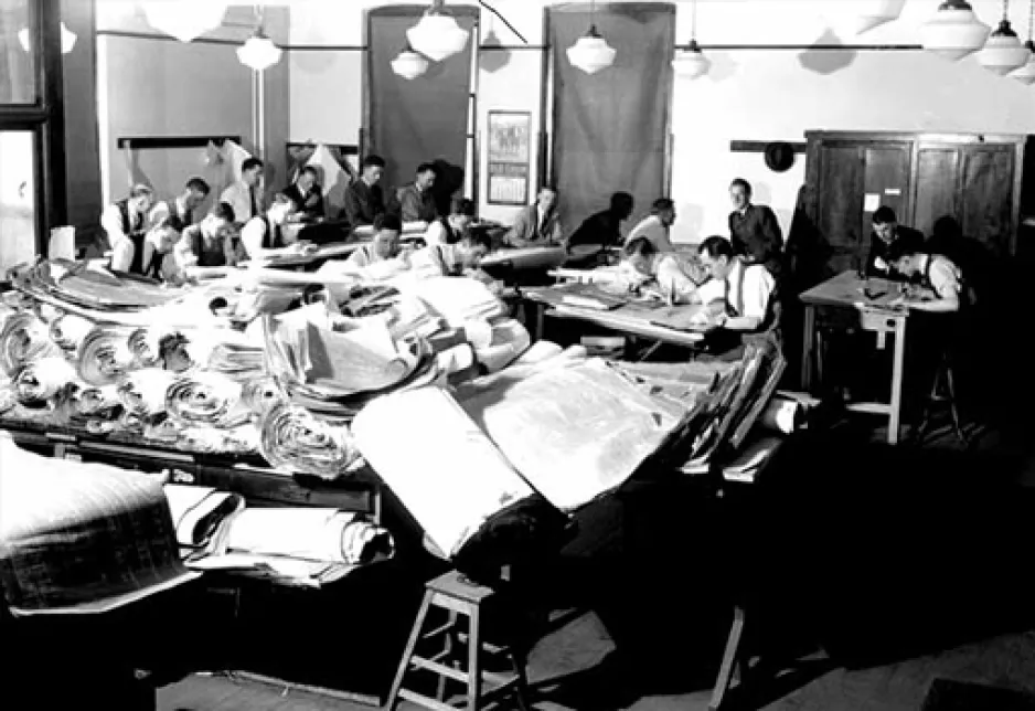 In a large room, men sit at tables working on building blueprints. In the foreground, a large table is heaped with blueprints — some sitting flat and others loosely rolled.