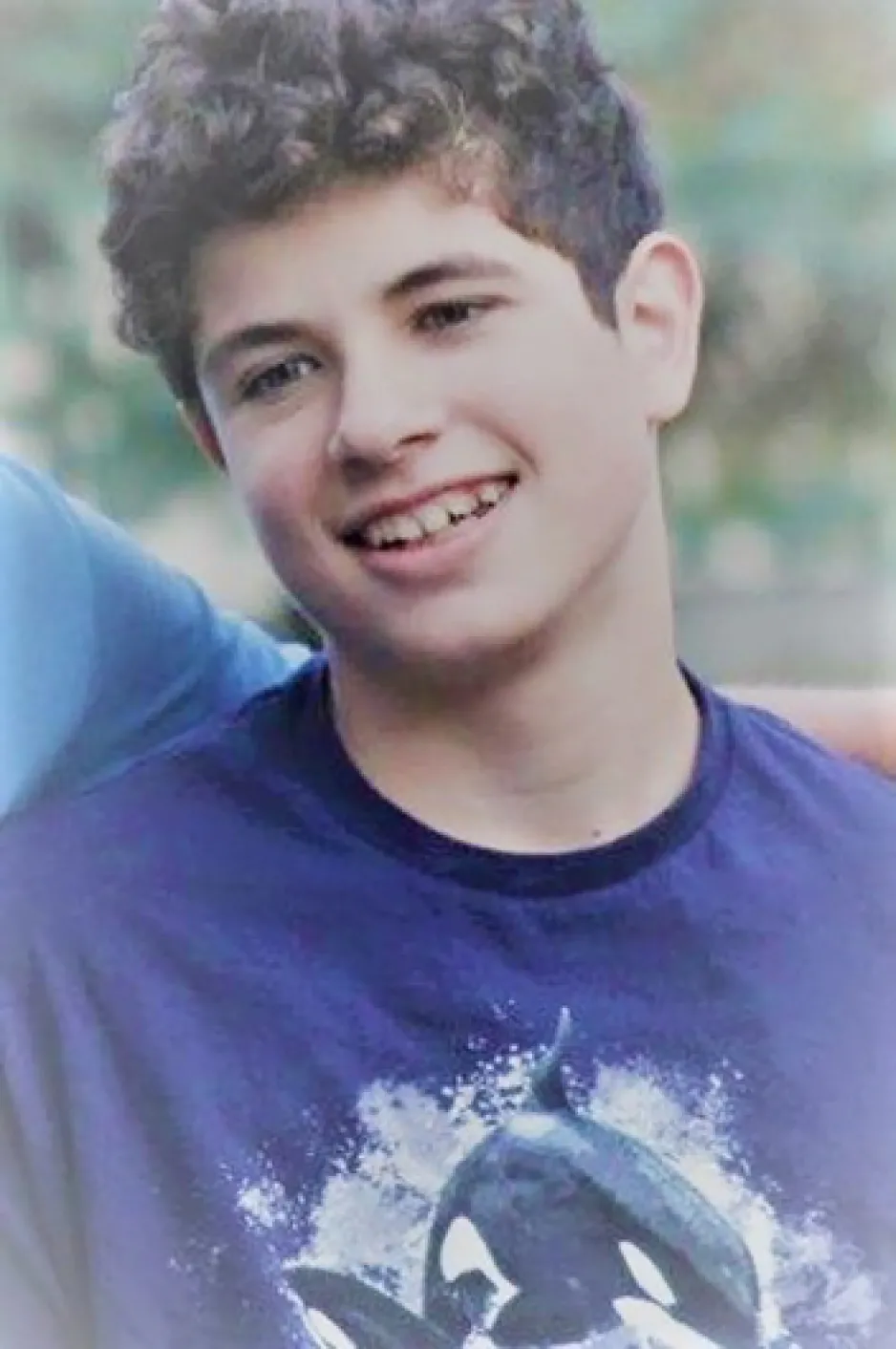 A teenaged boy wearing a purple shirt smiles and looks down from the camera.