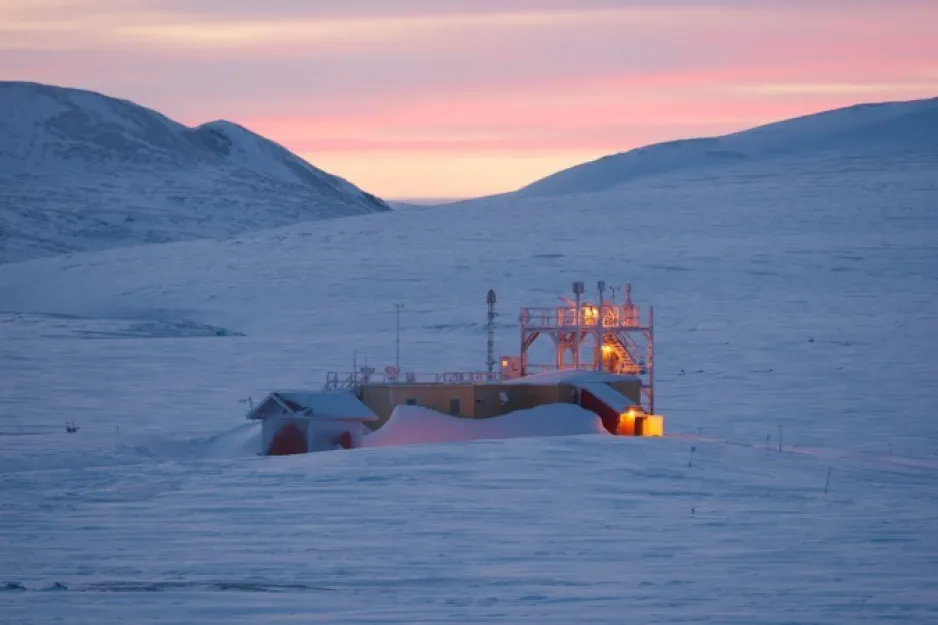 Un paysage stérile et enneigé sous un ciel rose, au coucher du soleil. On y voit une station de recherche utilisée par Environnement et Changement climatique Canada et ses halos de lumières.