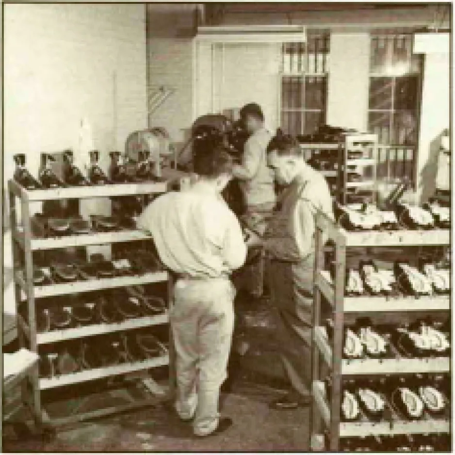 A black-and-white image shows three men are standing in a room filled with shelves of shoes. One of the men is working with a shoe press at the back of the room. Bars are visible in the windows.
