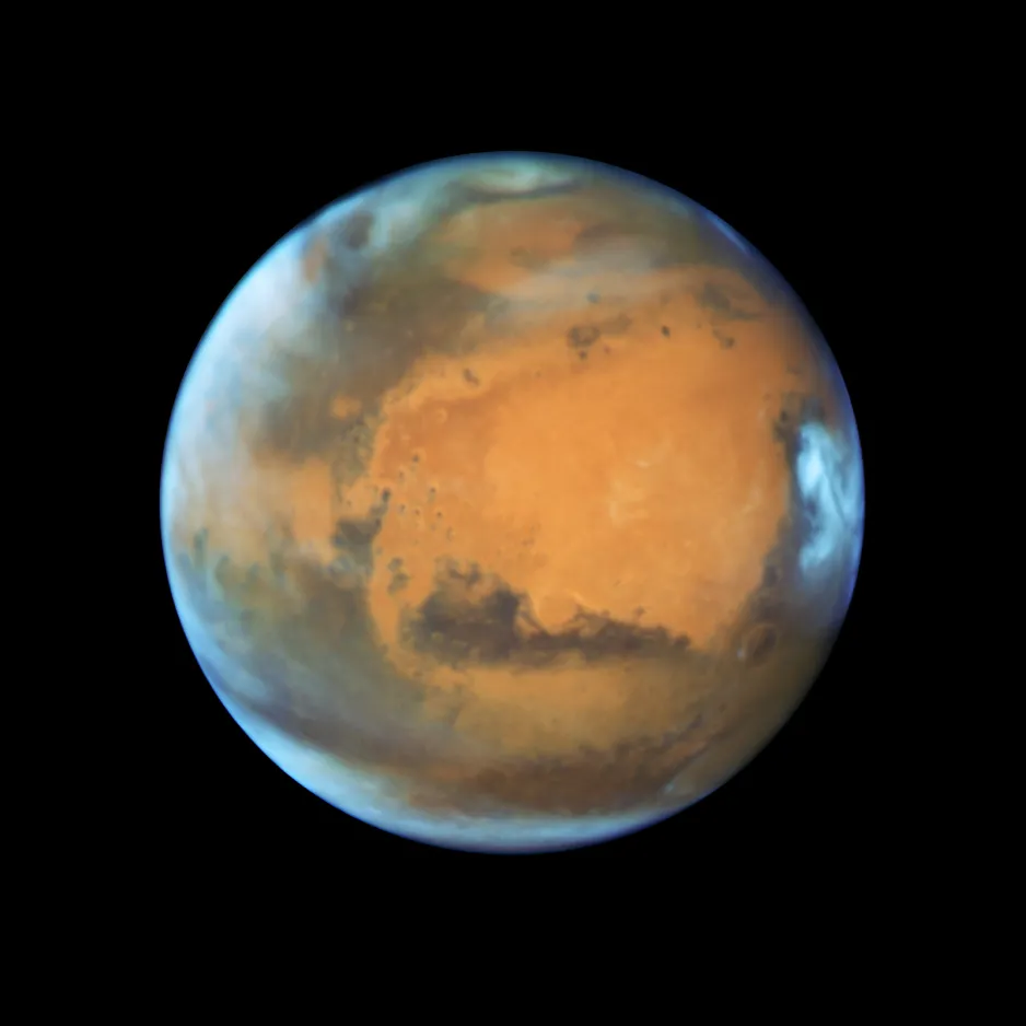 Image de Mars, captée par le télescope spatial Hubble. Elle montre la surface en grande partie rougeâtre et brune de la planète, et quelques nuages blancs et vaporeux. On peut y voir la calotte glaciaire au pôle Nord et quelques grands cratères.