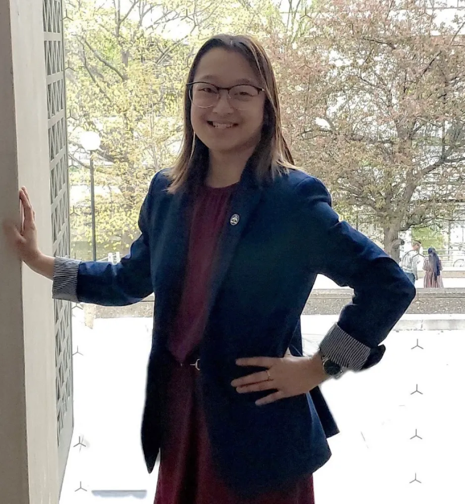 A young woman wearing glasses and a navy blue blazer smiles as she stands with one hand on her hip, and the other hand on the wall. She appears to be standing in front of a window; large trees are visible behind her.