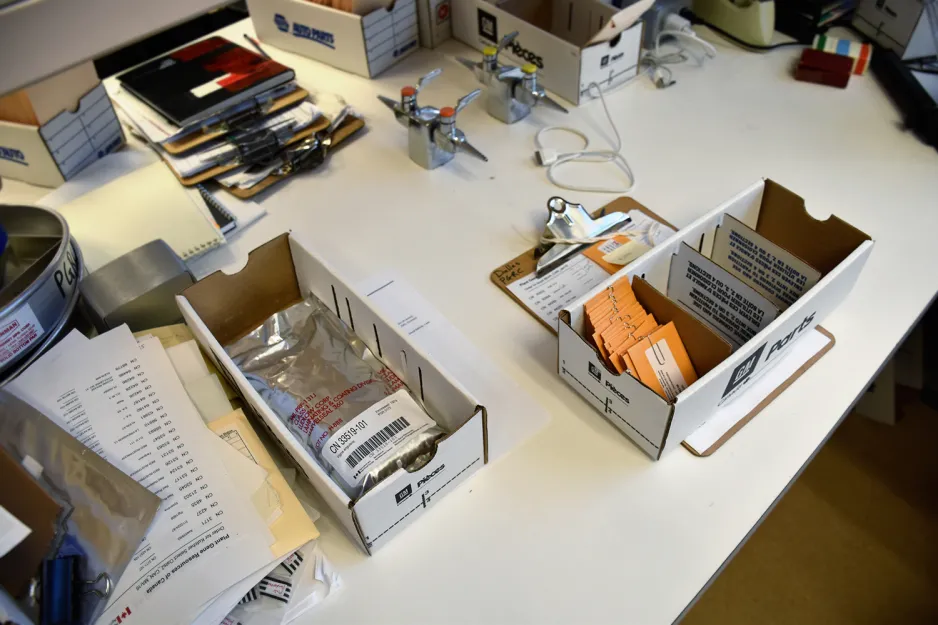 A photo of a white lab bench, where seed samples are being prepared for shipping. To the right, a box of small yellow envelopes containing seed samples sits on a clipboard holding a sheet of paper with a printed list on it. To the left, there’s a box containing a silver pouch with a barcode label.