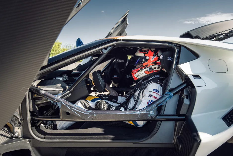 A driver wearing a white suit and a red-and-black helmet sits in the driver’s seat of a race car, with the doors open. The bars of the internal cage are visible, and another man can be seen leaning in on the passenger side of the car.