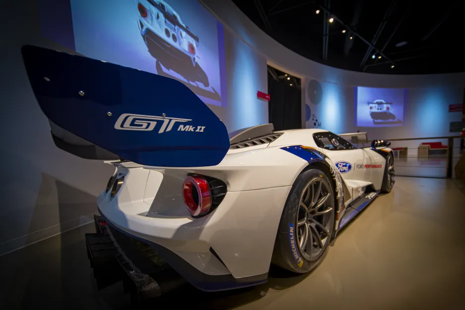 A white racecar with blue, yellow, and black accents sits inside a large indoor space. The image is taken looking from rear to front along the right-hand side of the car, with the camera right next to the large back spoiler. The letters “GT Mk II” are visible in white lettering on the blue spoiler. 