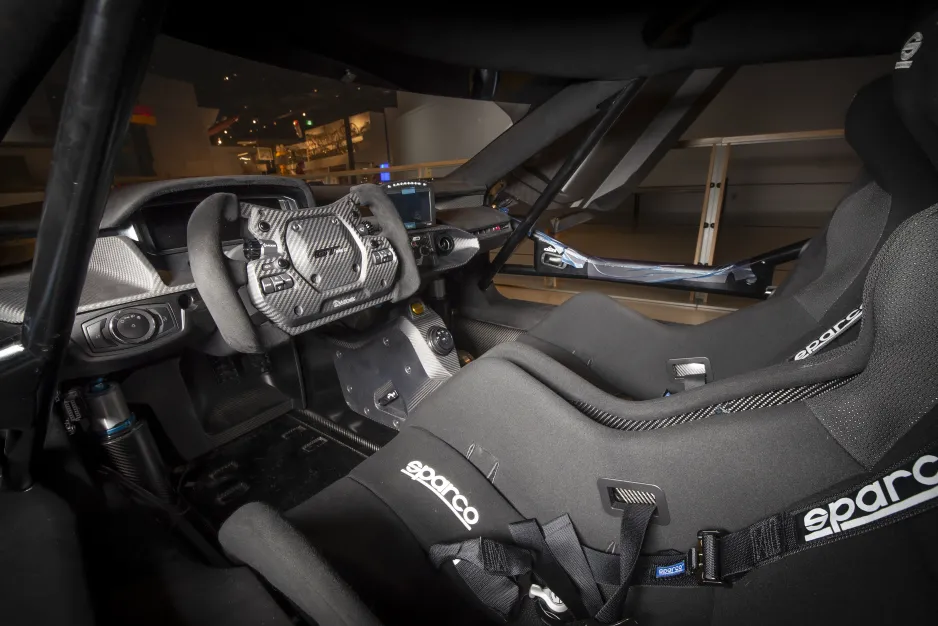 The dark interior of a racecar’s cockpit is pictured from the driver’s side. Both the driver and passenger seats have high sides, and seatbelts are visible. The steering wheel is elongated to almost a rectangular shape. Black metal bars form a cage inside the car.