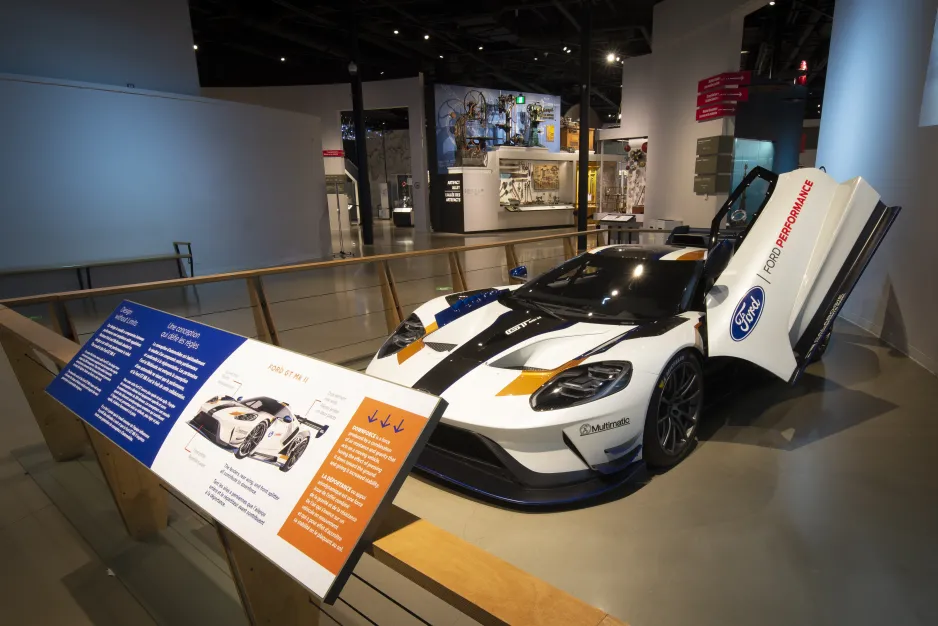 A white racecar with blue, yellow, and black accents sits inside a large indoor space behind an exhibition barrier. A museum panel covered in text is visible. The driver’s door opens vertically, and since it is propped open in the photo it is raised above the vehicle.   
