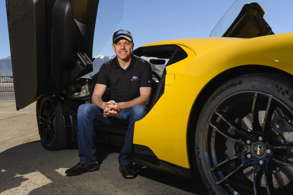 A man wearing jeans, a black shirt and a black baseball cap smiles as he sits on the edge of the driver’s seat of a bright yellow sports car. The sky is bright blue, and mountains are visible in the background.