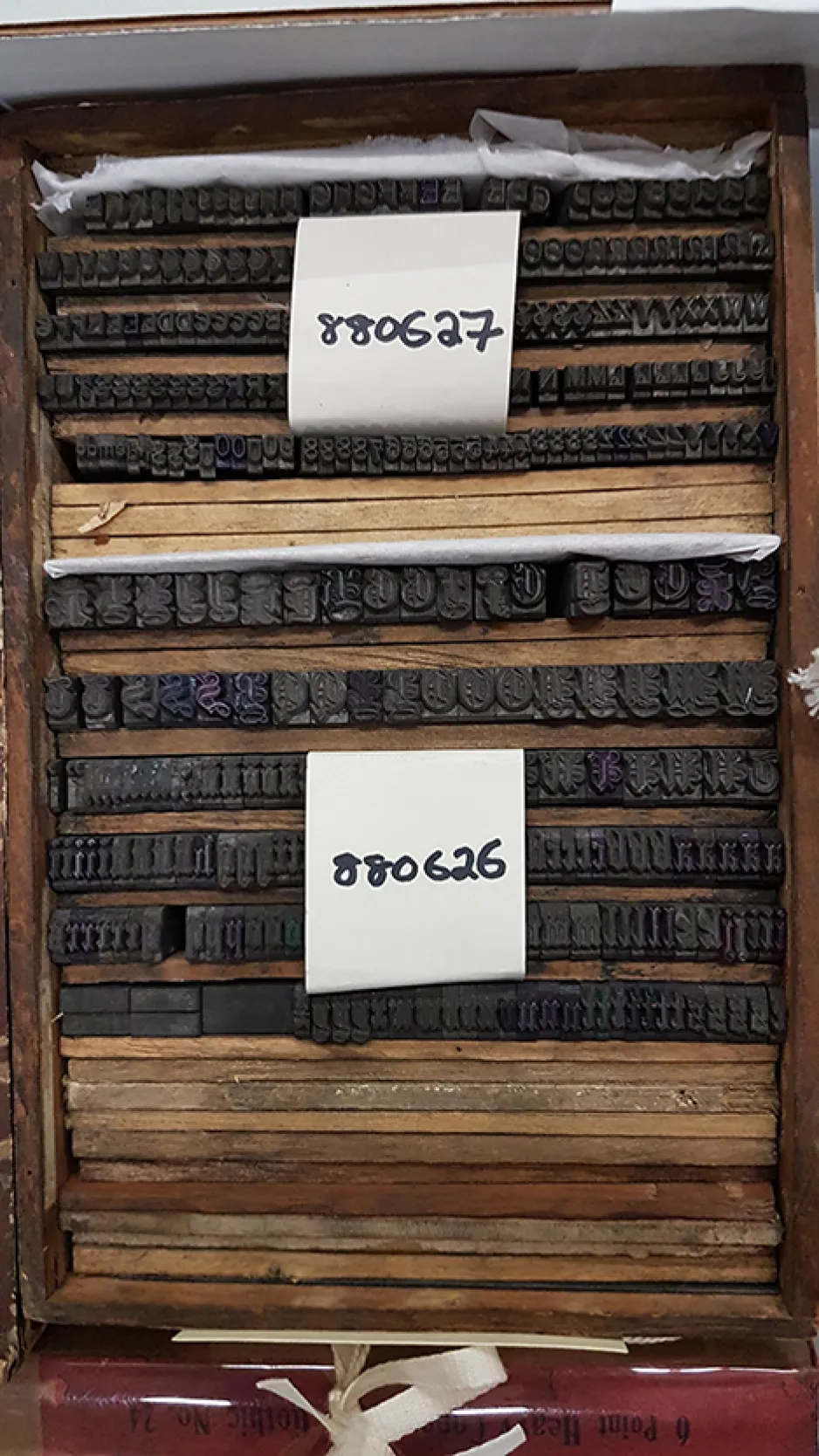 Two artifacts are housed in a wooden box. They are made up of foundry type that are organized alphabetically right to left. The first artifact is labeled with a white tag with black writing that says “880627.” The artifact is an 18-point ornate gothic typeface with serifs on the characters. The second artifact is labeled with a white tag with black writing that says “880626.” It is a 24-point Black script with very ornate characters.