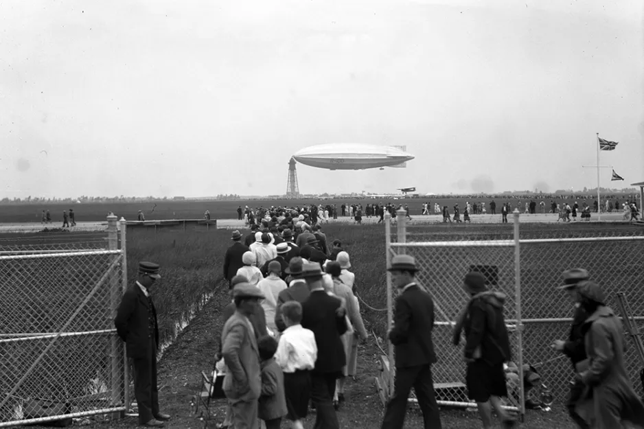 Une photographie noir et blanc montre une file de personnes passant par l’entrée d’une barrière clôturée et, en arrière-plan, un immense dirigeable amarré au sommet d’une haute tour qui ressemble à un phare.