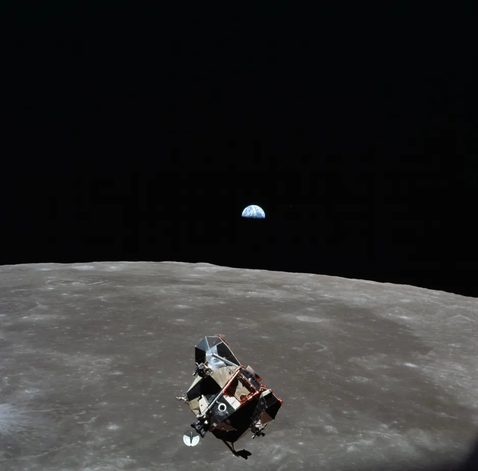 View of the Apollo 11 ascent module in the foreground flying above the grey Moon. Above the Moon’s horizon, the Earth is partly in the Moon’s shadow against the black of space (known as Earthrise).