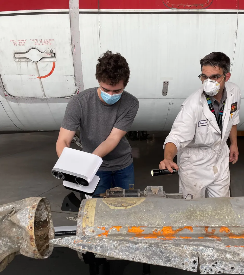 Two men wearing COVID-19 masks stand behind a dented aircraft model that is grey with orange rust. The man on the left holds a white, 3D scanner up to the artifact, while the man on the right holds a flashlight.