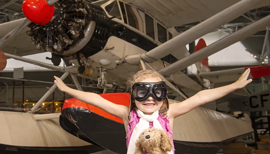 A young girl wearing aviator goggles, a white scarf and a brown aviator helmet sits in front of a float plane in the museum. She has a brown stuffed dog on her lap and her arms are outstretched as though she is flying. 