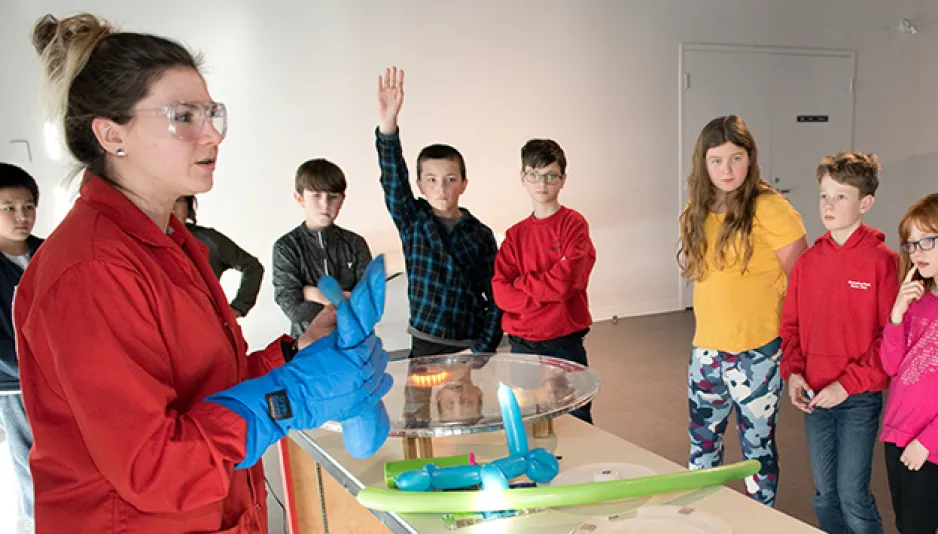 A person in a red lab coat, protective glasses and blue gloves is standing in front of a table with various objects on it. There is a large group of children standing in front of the table watching, and two of the children have their hands raised in the air.  