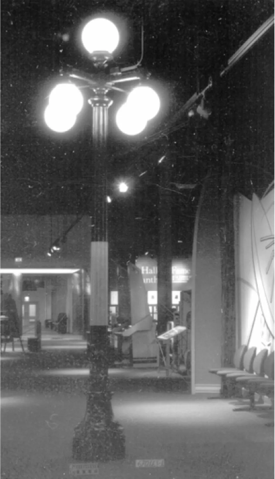 Black and white image of an old streetlamp topped with five brightly glowing globes. The lamp post is displayed indoors in a museum setting.