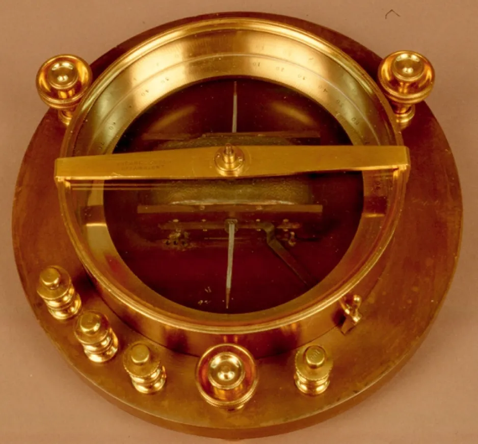 Brass-coloured ring-shaped device mounted on a slightly larger-diameter round, wooden base. The device has a glass front and has seven small brass posts around its perimeter.