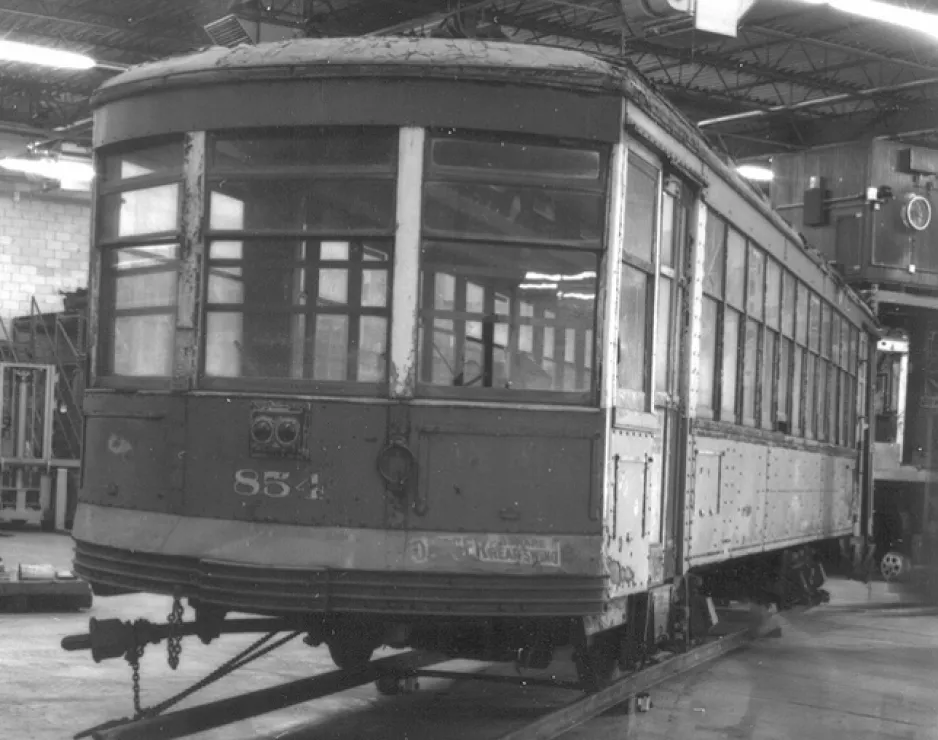 A black and white photo of an old streetcar showing number 854 on its front. The streetcar has windows on all sides and sits on temporary streetcar tracks in a warehouse.