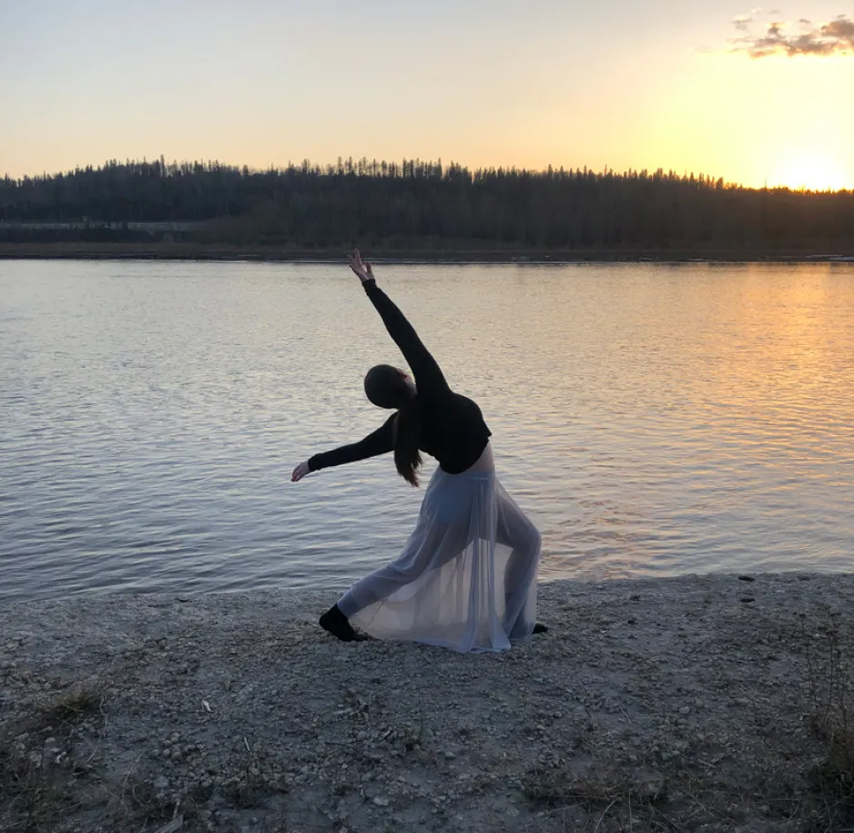 A woman in a dark shirt with a translucent flowy skirt strikes a dance pose next to a river. The sun is low on the horizon.