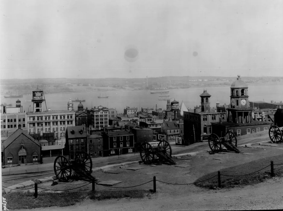 A black and white photograph showing four cannons in the foreground. Mid-ground shows buildings and houses, and the background is a large body of water dotted with ships.