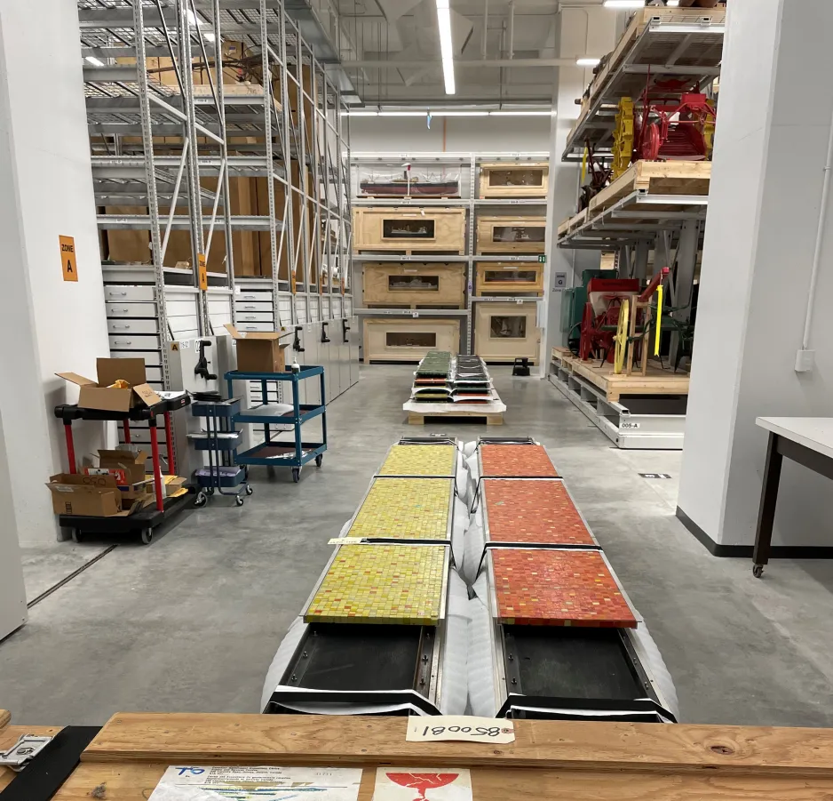 Wooden pallets carrying yellow, orange, and green mosaic-tiled panels in the middle of the aisle of a large storage room with metal shelving holding artifacts. 