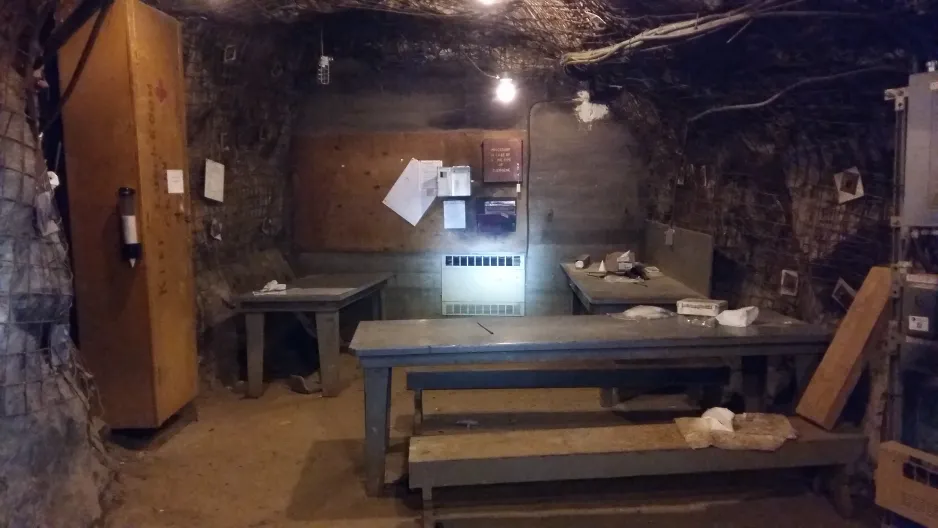 Une chambre de refuge souterraine, avec des tables et des bancs. L’espace est faiblement éclairé par plusieurs petites ampoules. 