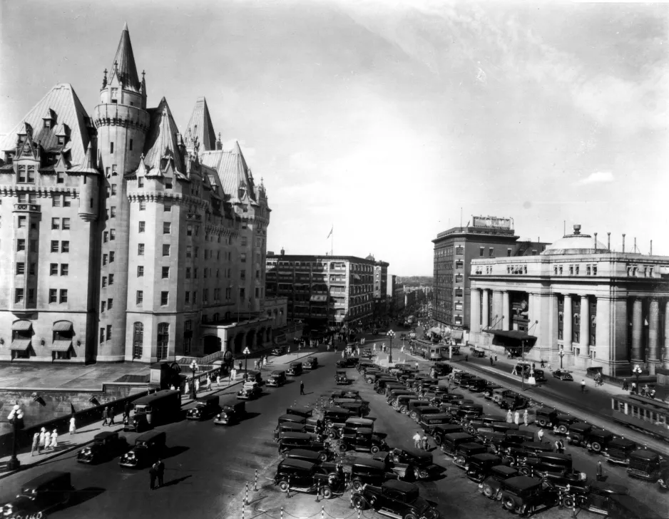 A black and white photograph of a castle-like stone building on the left-hand side of a road, and a smaller stone building with columns and a dome roof on the right-hand side. The road has many black, 1930s -style cars parked in the centre lane.