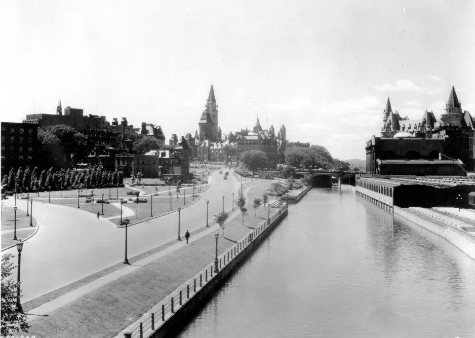 Black and white image showing a canal in the centre, flanked on the left side by a street, walking path, and park, and on the right side by railway tracks. The spires of a large, stone building can be seen in the distance.