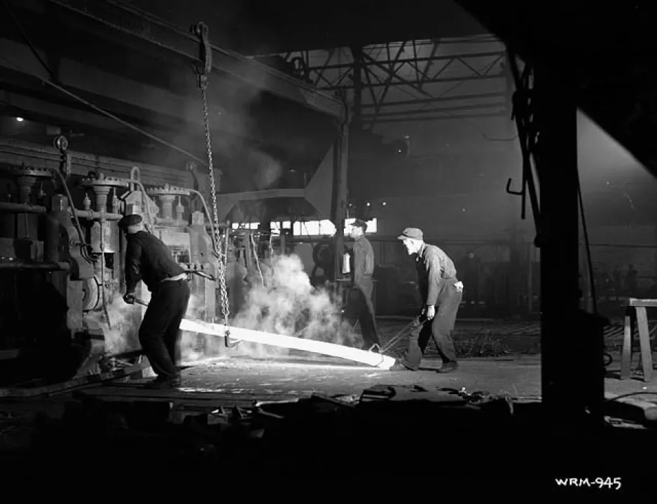Une image noir et blanc montre deux ouvriers en train d’opérer un laminoir dans un bâtiment industriel.