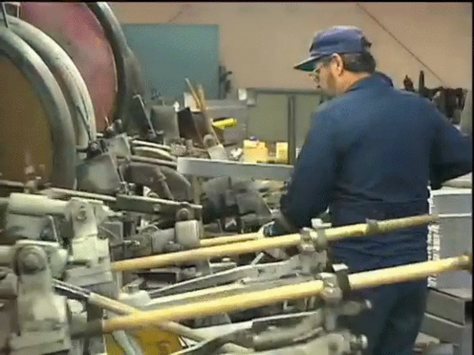 Un ouvrier actionne une machine à clous; l’image montre une imposante machinerie dans ce qui ressemble à un entrepôt.