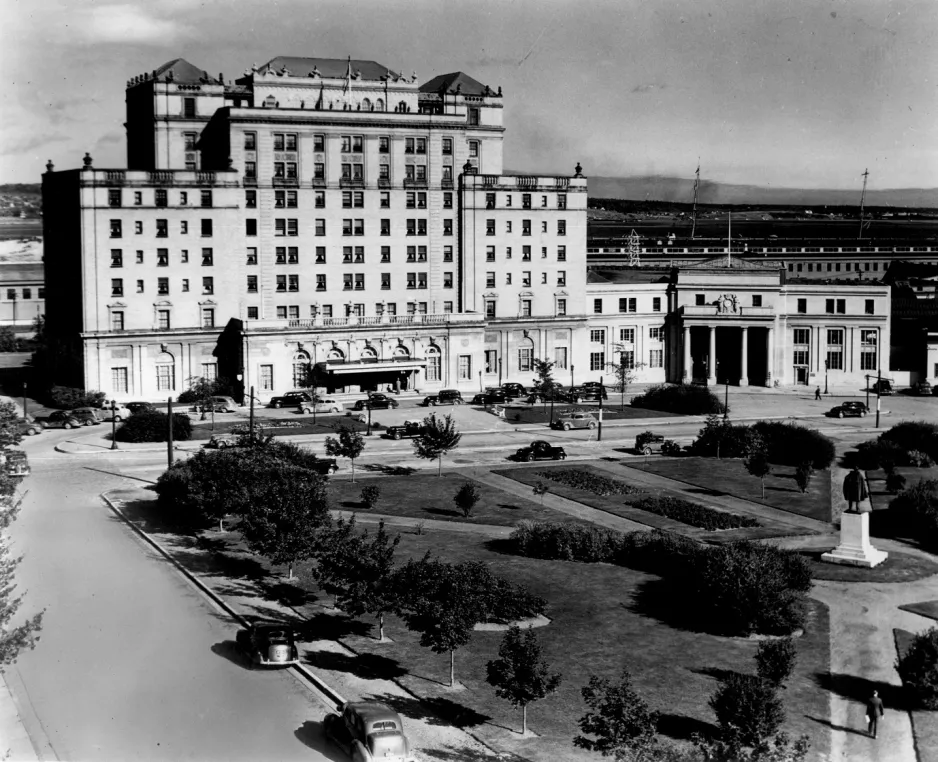 A black and white photograph featuring a nine-story, ornamental building with a round driveway in the front. A green lawn with statues and walkways is in the foreground.