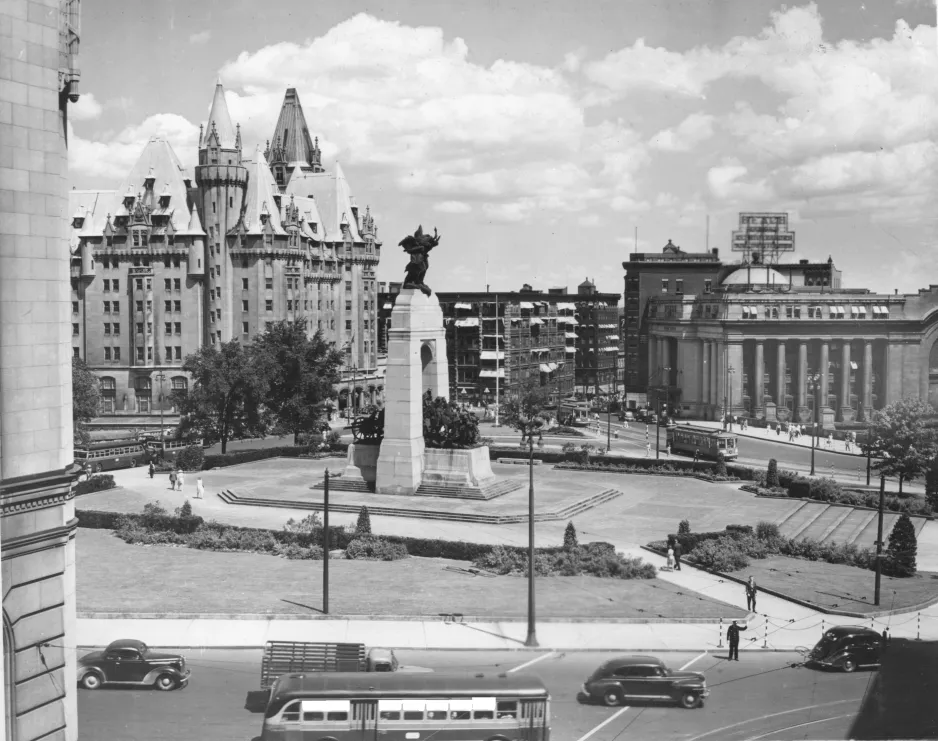 A black and white photograph with a road in the foreground, a tall stone and bronze monument in the mid-ground, and a castle-like stone building in the background.