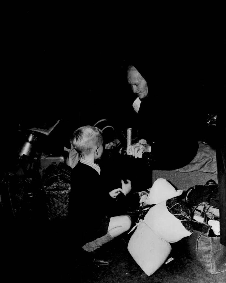 A black and white photograph of an elderly women wearing black, and a small boy sitting beside her. Luggage surrounds them.