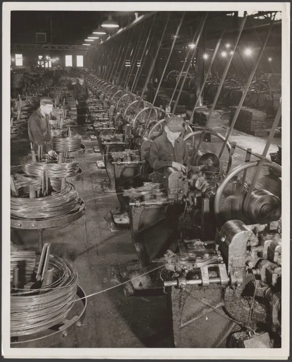 Une image noir et blanc montre des ouvriers qui opèrent des machines servant à la fabrication de clous de Paris.