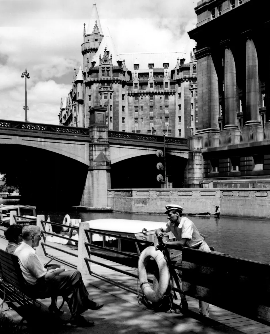 A black and white photograph of a man and woman sitting on a bench beside a canal, talking to a man wearing a sailor hat, standing beside a small boat. In the background there is a bridge, and the spires of an ornate stone building.