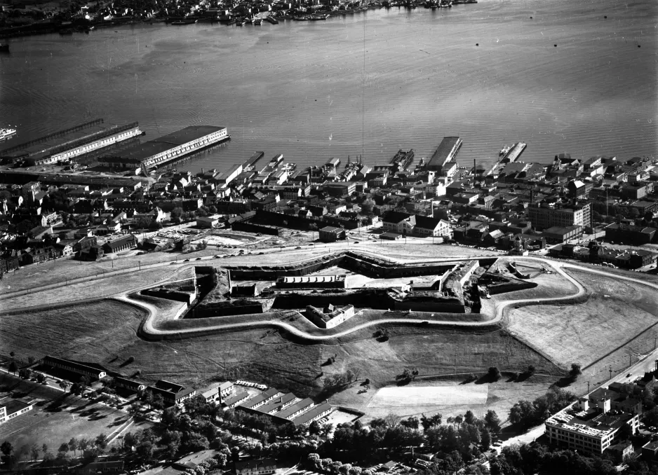 A black and white photograph taken from a high vantage point, with a cluster of long buildings surrounded by a star-shaped stone wall. In the background, houses and buildings stretch towards a harbour. Docks and large buildings used for immigration processing jut into the water.