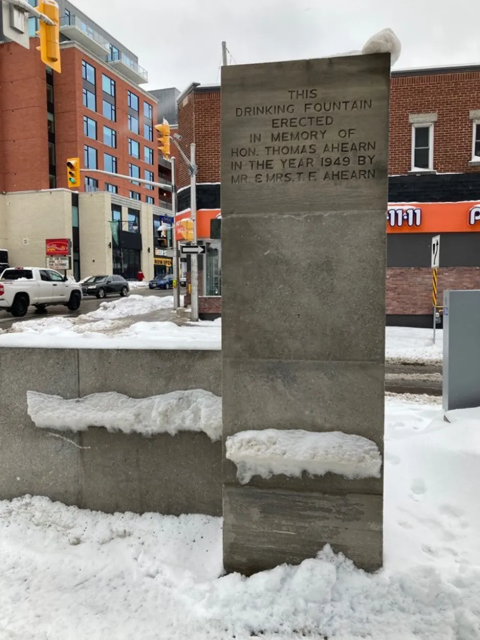 A tall grey stone rectangle is covered in snow. Writing etched into the top reads: This drinking fountain erected in memory of Hon. Thomas Ahearn in the year 1949 by Mr & Mrs T.F. Ahearn. 