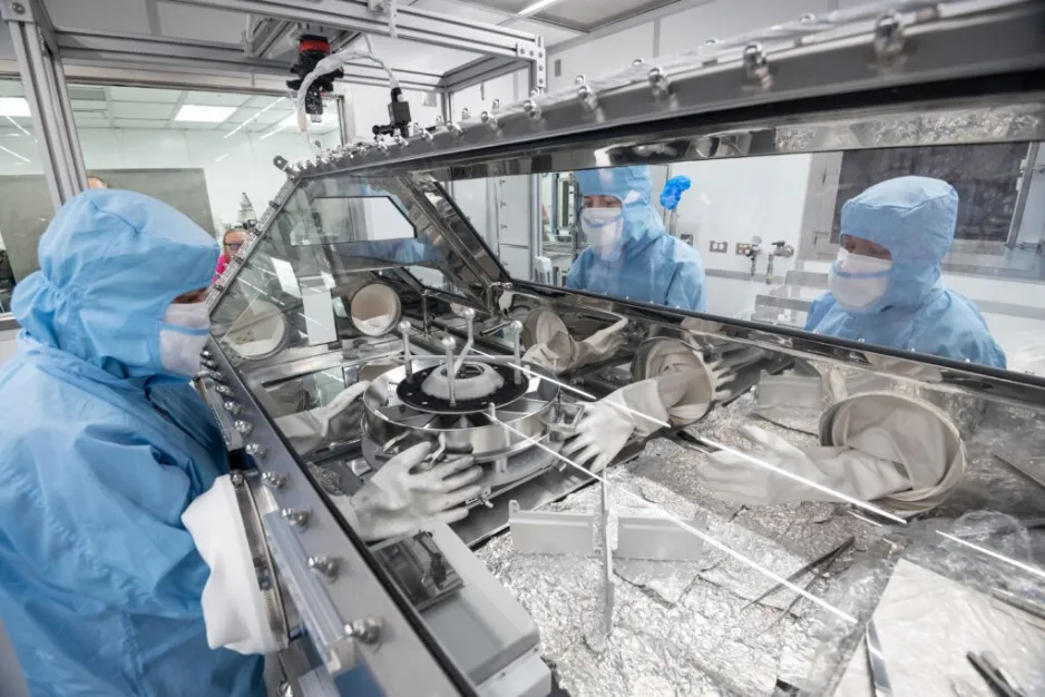 : Trois personnes vêtues de combinaisons de laboratoire bleues interagissent avec un grand contenant d’échantillons se trouvant dans un espace de travail vitré et scellé hermétiquement.