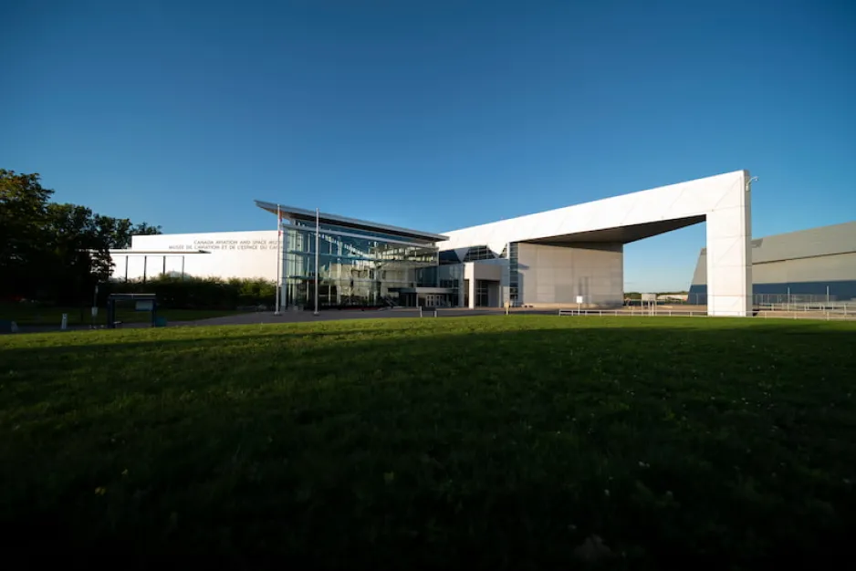 The angular façade of a large, white building, with the words “Canada Aviation and Space Museum” visible on its left side. Green grass is visible in the foreground and a clear blue sky in the background.