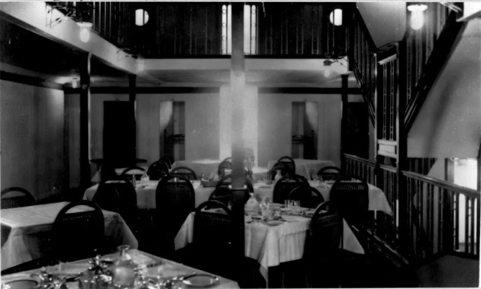 Une photographie noir et blanc montre des tables dressées en vue d’un élégant repas, avec vaisselle en porcelaine.