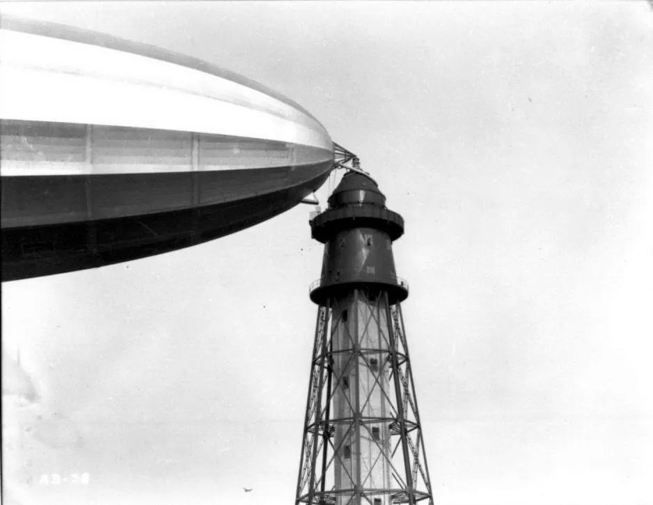 Une photographie noir et blanc montre en gros plan le nez d’un immense dirigeable, amarré au sommet d’une haute tour qui ressemble à un phare.