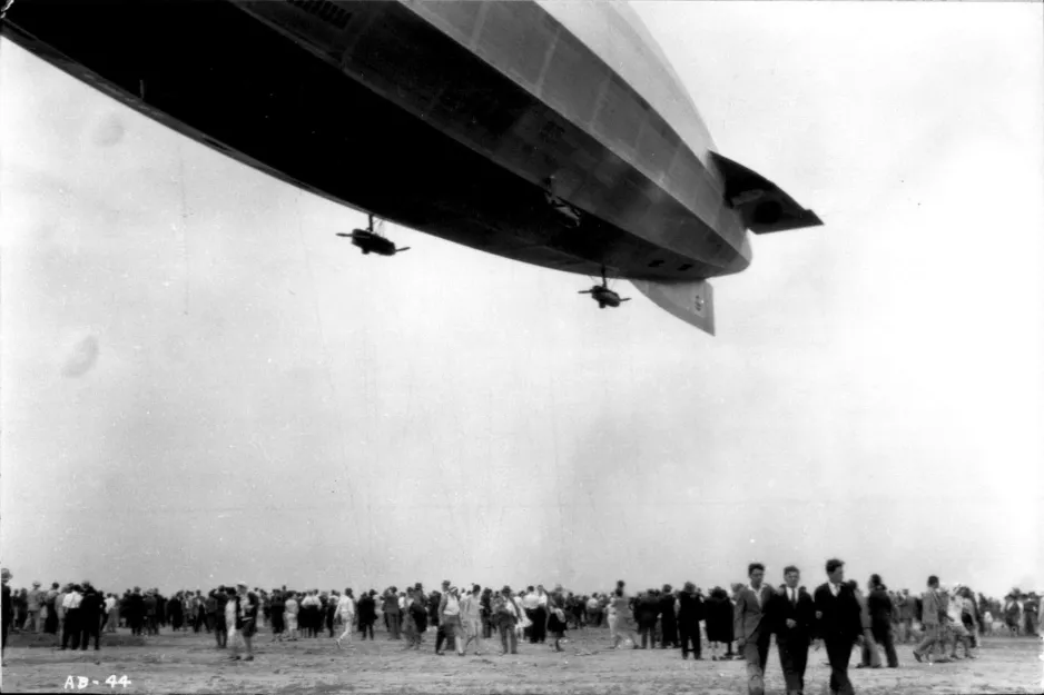 Une photographie noir et blanc montre la moitié arrière d’un immense dirigeable volant au-dessus d’une foule. Deux petites nacelles moteurs sont fixées au ventre du dirigeable.