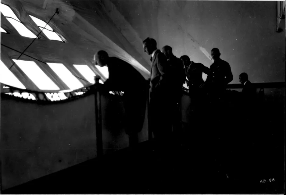 Une photographie noir et blanc montre, à l’intérieur du R-100, les silhouettes de six personnes qui regardent vers l’extérieur à travers un ensemble de petites fenêtres carrées.