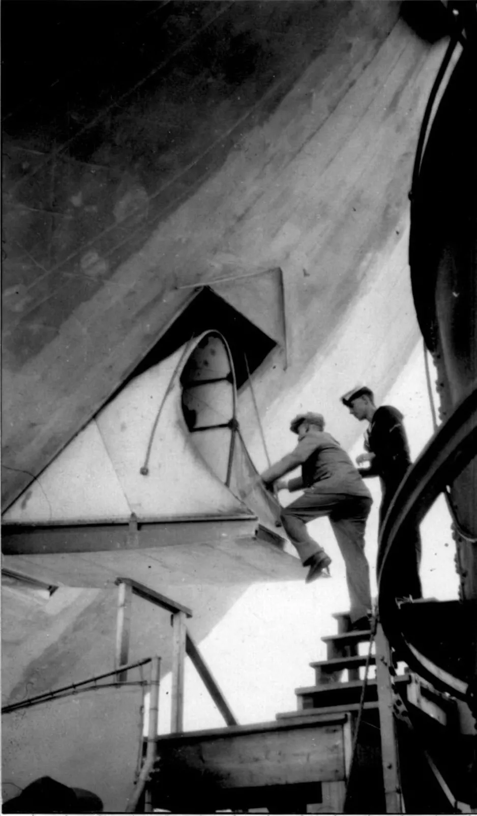 Une photographie noir et blanc montre un homme juché sur un escalier, en train de se hisser à bord du R-100. La hauteur à franchir est d’environ 1 m. Un autre homme se tient derrière lui.